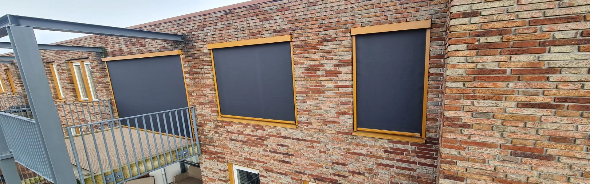 Gebouw met bakstenen gevel, voorzien van zonwering in antraciet kleur met goudkleurige frames