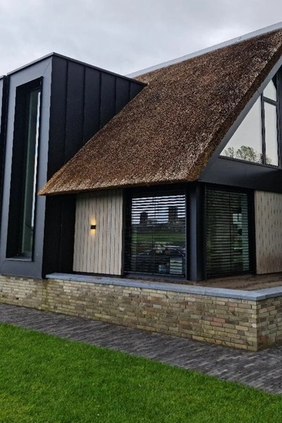 Buitenjaloezie raffstore bij zijgevel van woning met rieten dak en grote glaspartij Buitenjaloezie raffstore bij zijgevel van woning met rieten dak en grote glaspartij