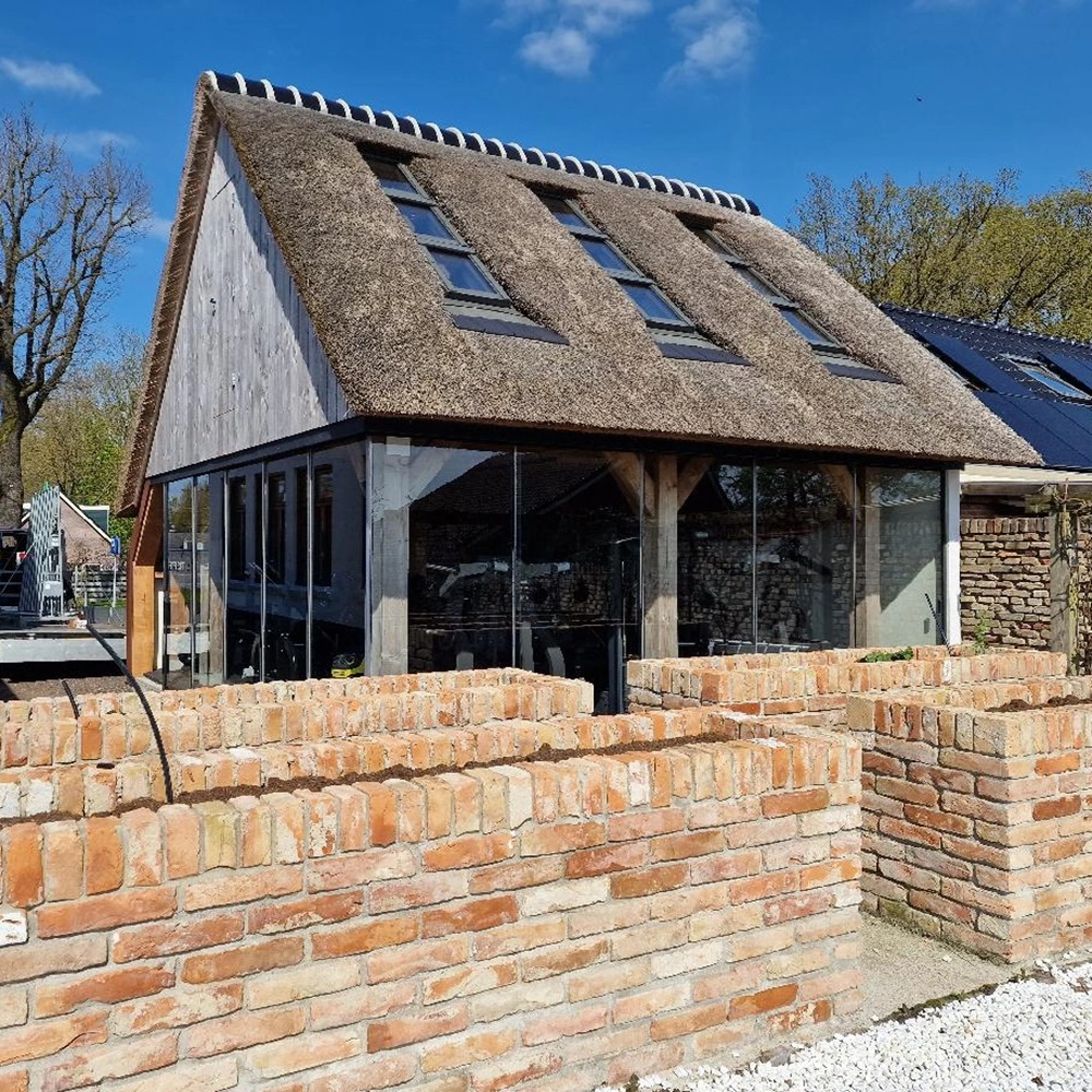 Houten bijgebouw met glazen schuifwand en rieten dak naast bakstenen muur