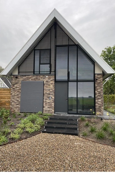 Louvre zonwering op gevel van huis met puntdak en grote ramen, aangelegd met grindpad en tuin Louvre zonwering op gevel van huis met puntdak en grote ramen, aangelegd met grindpad en tuin