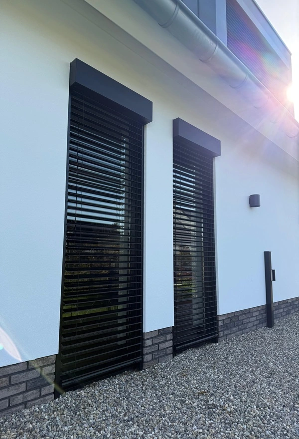 Twee smalle zwarte buitenjaloezieen tegen een witte muur, met grind aan de onderzijde Twee smalle zwarte buitenjaloezieen tegen een witte muur, met grind aan de onderzijde