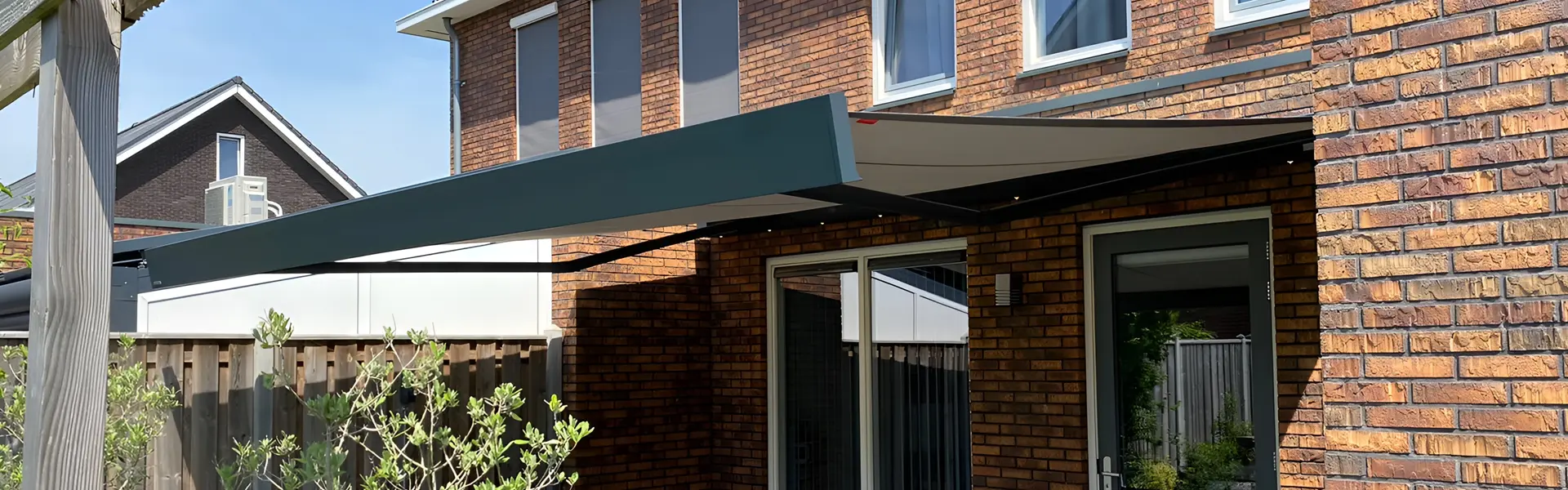 Donkergrijs knikarmscherm gemonteerd aan een bakstenen gevel, met een moderne buitenruimte