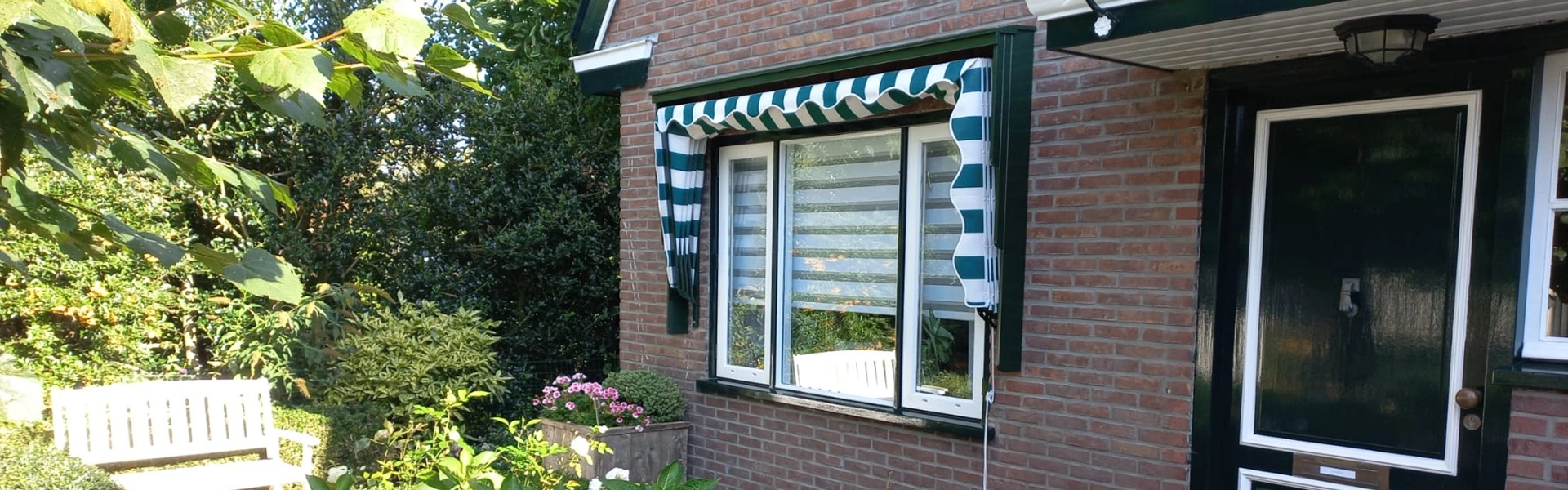 Groene en witte gestreepte markies bij het raam van een bakstenen huis met bloementuin