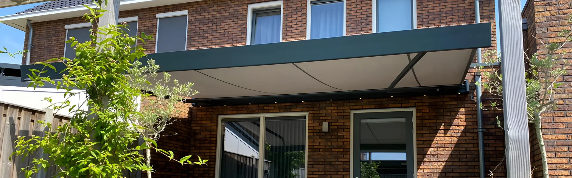 Knikarmscherm met donkere doekkleur boven een zonnige patio en groene tuin