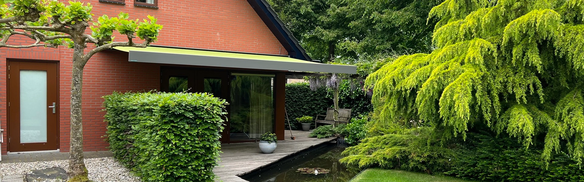 Kubata knikarmscherm bij rode bakstenen woning met groene tuin en vijver