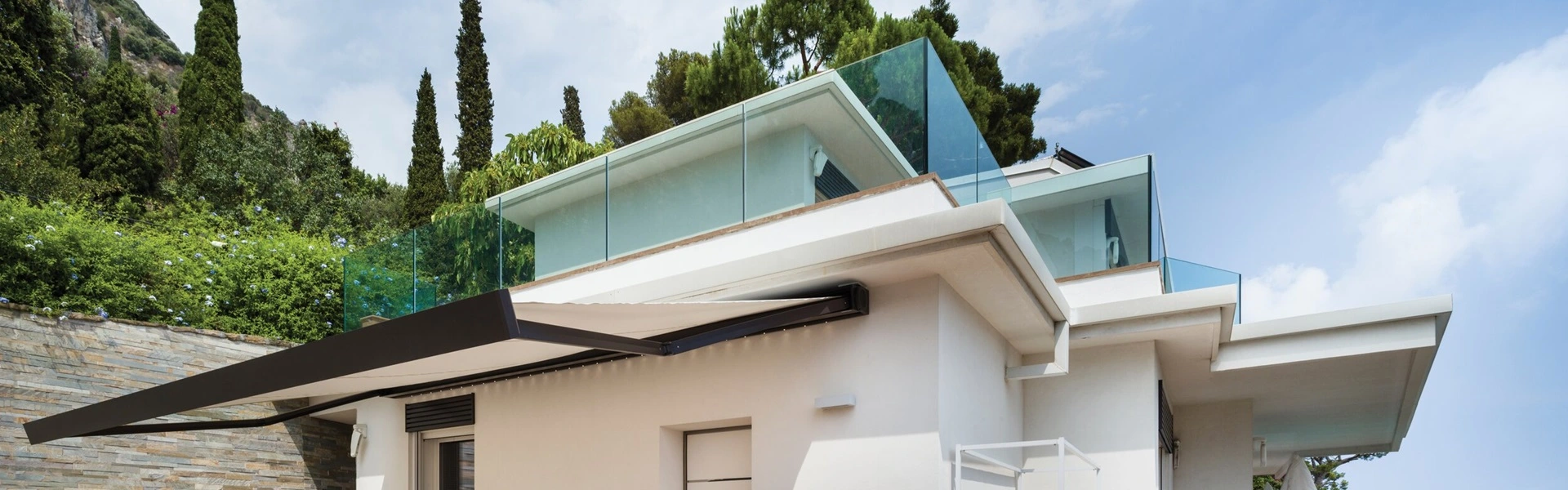 Nodo knikarmscherm over terras van huis met glazen balustrade en groene omgeving