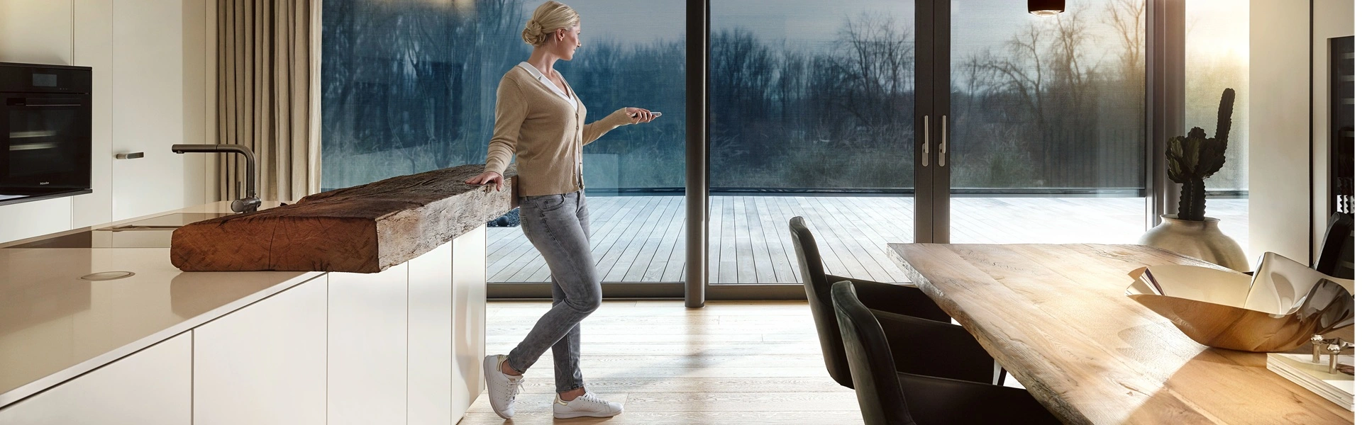 Vrouw bedient domotica in keuken met houten details en uitzicht op terras