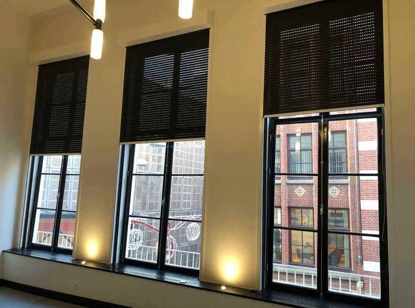 Zwarte rolluiken in een moderne ruimte met hoge ramen en uitzicht op stadsgebouwen